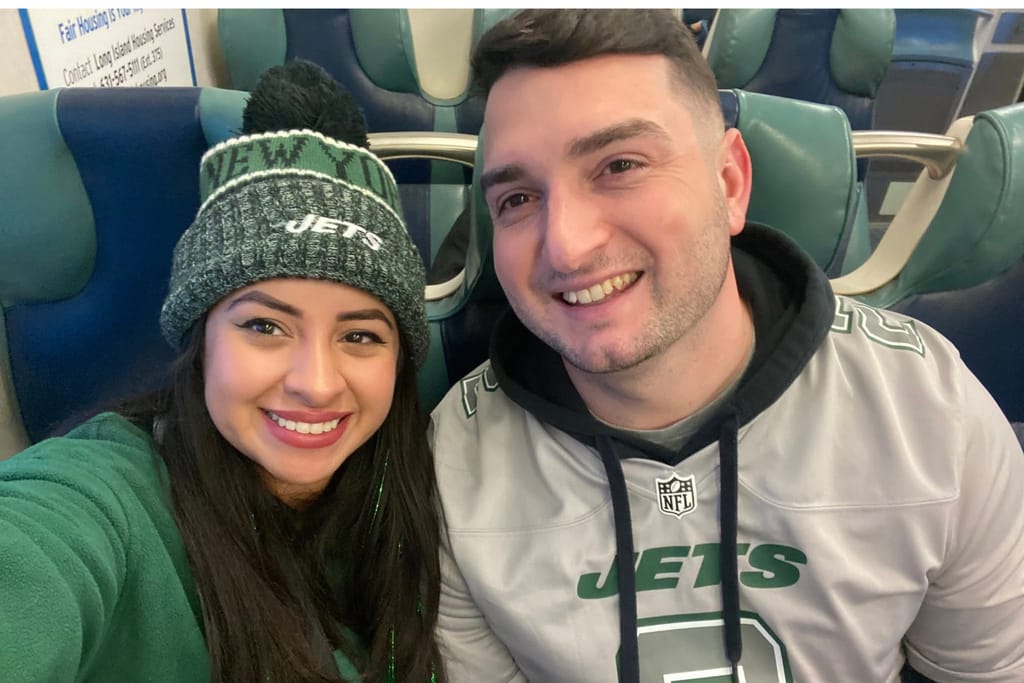 Carolyn Infantas and Chris Durham smiling and seated on an LIRR train. They are wearing New York Jets gear.